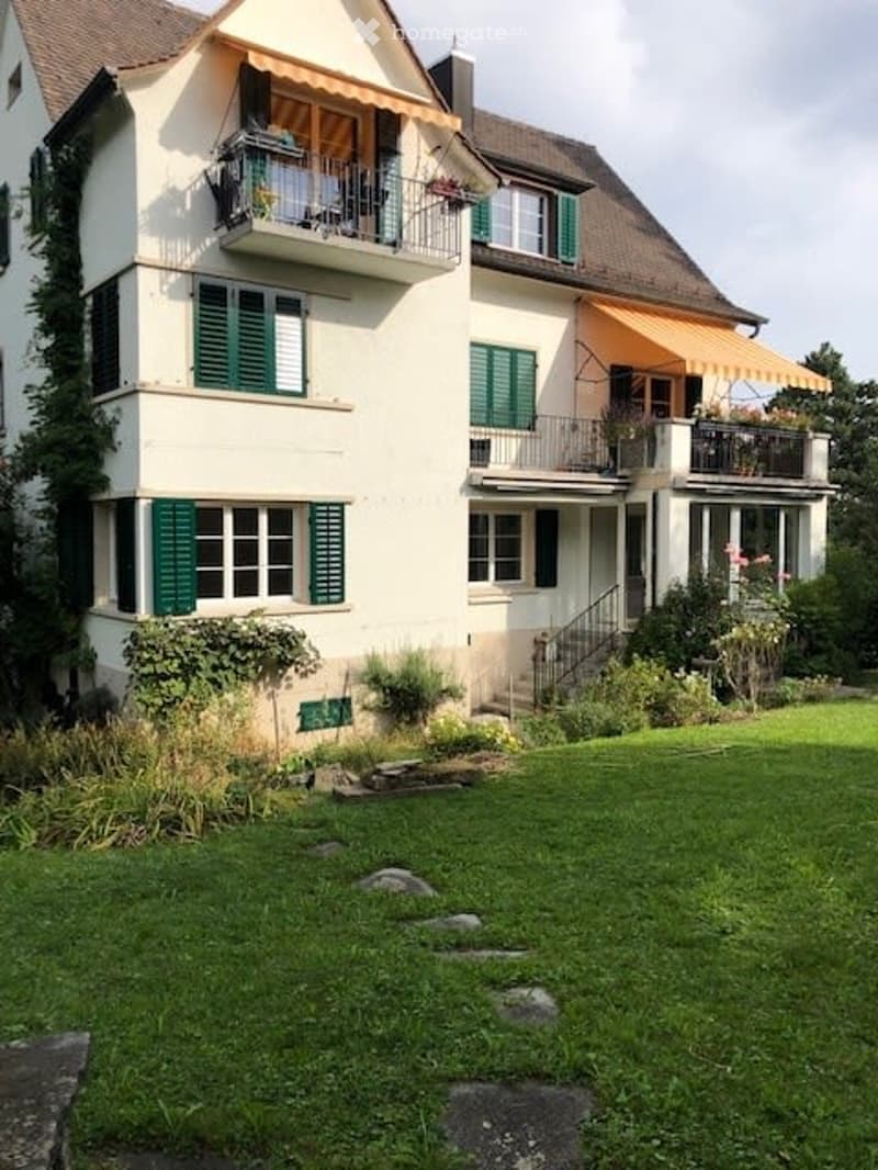 A two-story house with white walls, green shutters, and multiple balconies. The house features a well-maintained garden with a lawn and some plants.