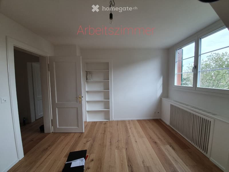 Empty room with wooden flooring, white walls, a built-in shelf unit, and large windows letting in natural light.