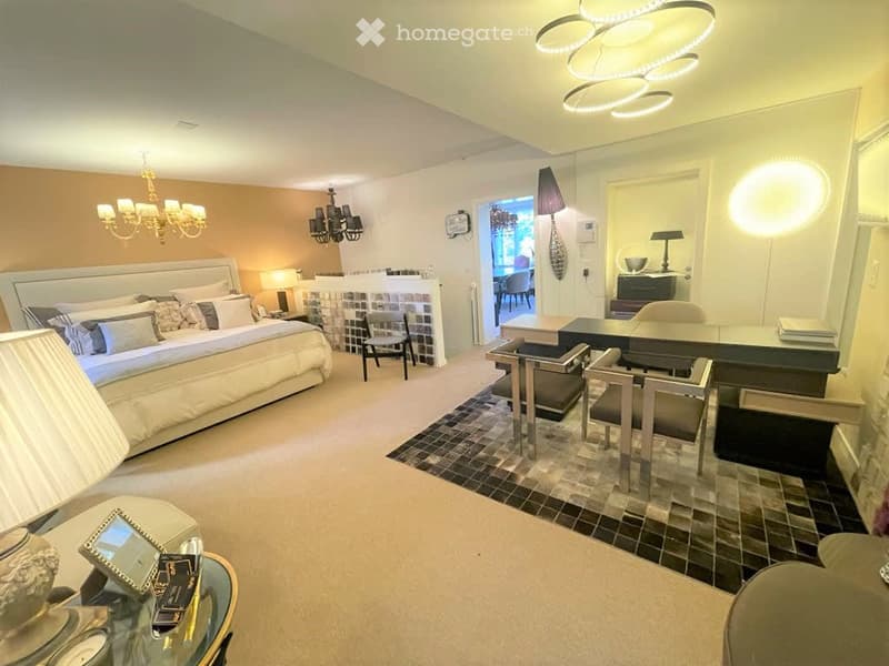 Elegant bedroom with a large bed, fireplace, nightstand, and a sitting area with chairs and a table. The room features modern lighting and a cozy ambiance.