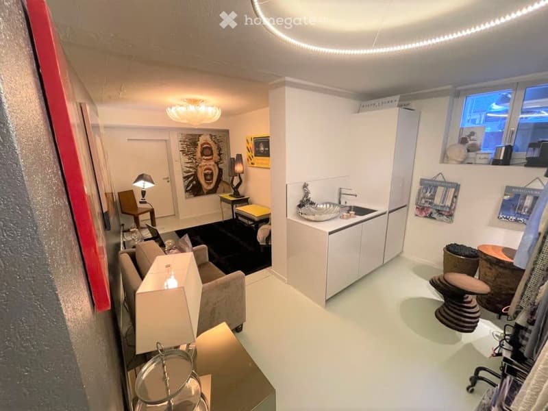 A modern, well-lit room with a sofa, small table, chairs, lamps, a kitchen area with cabinets and sink, and decorative items including a picture frame and stools.