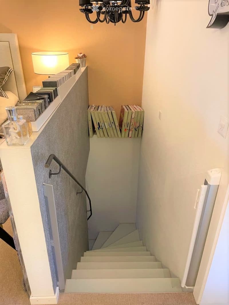 Narrow staircase with white steps and a modern railing, flanked by light-colored walls and a chandelier hanging above. A small shelf on the left holds books, a lamp, and decorative items.