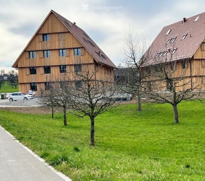Two-story wooden houses with large windows and pitched roofs, surrounded by a grassy area with trees and a parking lot.