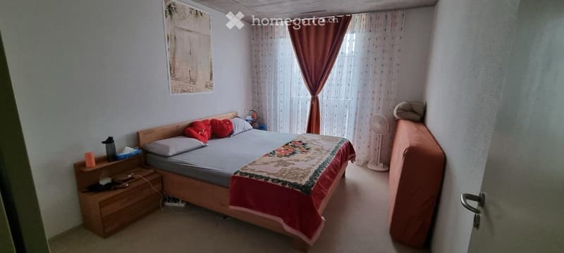 Cozy bedroom with a double bed, nightstand, fan, and a picture frame on the wall. The bed is made with pillows and a colorful blanket.