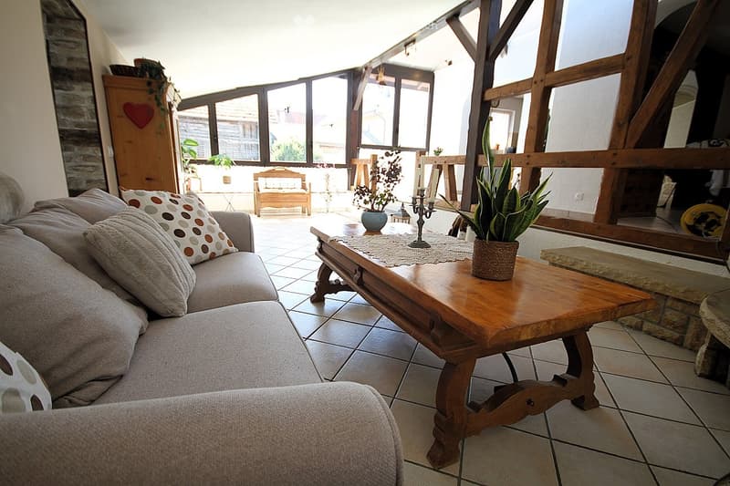 Bright living room with large windows offering a view, featuring a beige sofa with patterned cushions, a wooden coffee table with a table runner, candle holder, potted plants, and a wooden bench in the background.