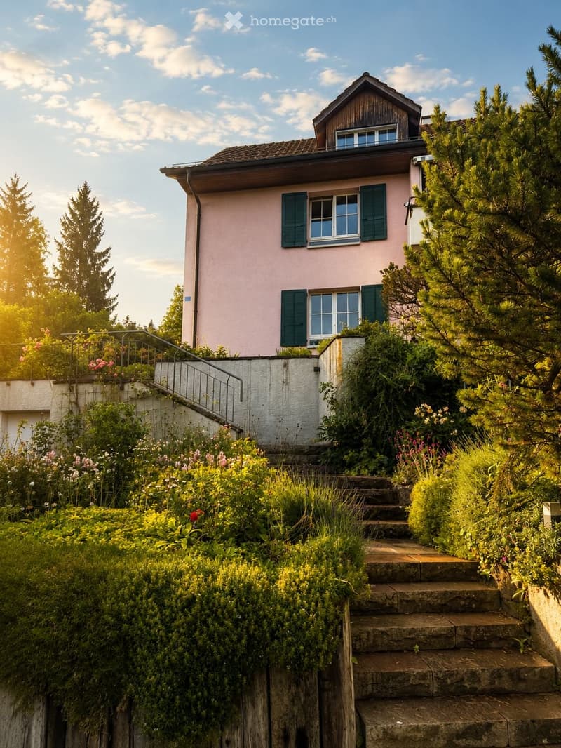 A two-story pink house with green shutters, surrounded by lush greenery and a stone staircase leading up to the entrance.