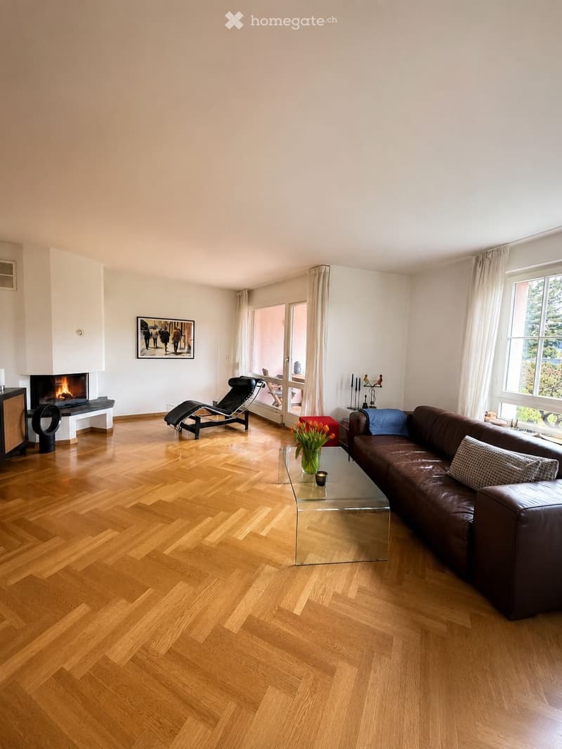 Modern living room with wooden herringbone flooring, a brown leather sofa, a glass coffee table, a fireplace, a lounge chair, and a vase with flowers on the coffee table.