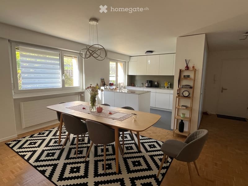 Bright, modern dining area with a wooden dining table, chairs, a vase with flowers, and a shelf with decorative items. The room features white cabinets, a chandelier, and large windows with blinds.