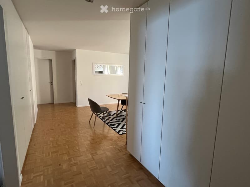 A small, modern room with wooden flooring, a single chair, and a small table. The room has white walls, a large mirror, and a patterned rug.