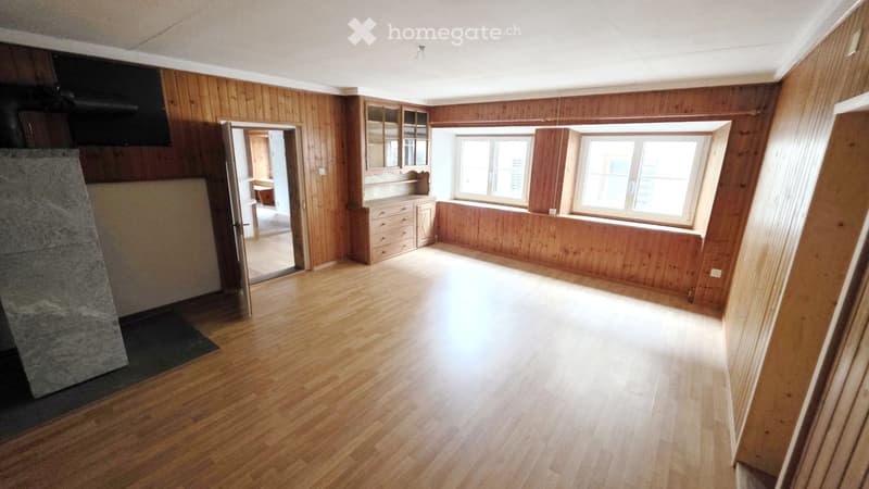 Empty room with wooden walls and floor, large windows, fireplace, and open doorway to another room.