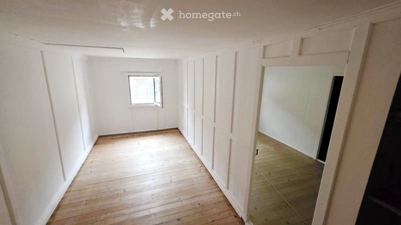 Empty hallway with wooden flooring and white walls, featuring a small window and a mirror on one side.