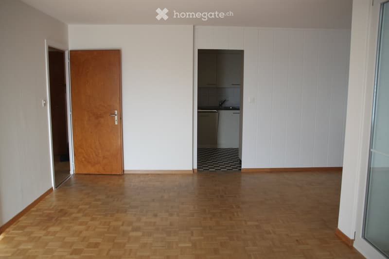 Empty room with wooden flooring, white walls, and a doorway leading to a small kitchen area with white cabinets and a checkered floor.