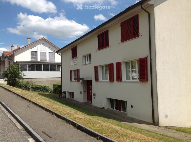 Two-story house with white walls, red shutters, and a sloped roof. The house has multiple windows and a small front yard with a bench.