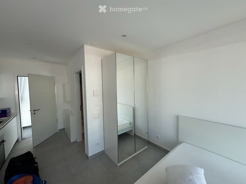 Modern, minimalist bedroom with a single bed, large mirrored wardrobe, and tiled floor. The room features white walls and a neutral color palette.