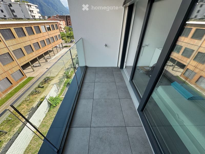 Modern balcony with large glass railings, tiled floor, overlooking a landscaped area with wooden structures and greenery.