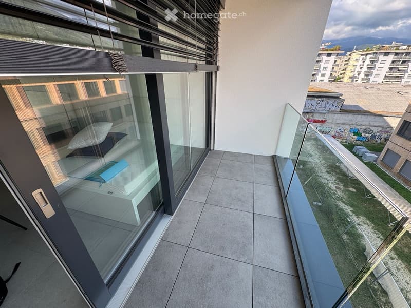 Modern balcony with large glass windows, tiled floor, and a view of the surrounding area with buildings and greenery.