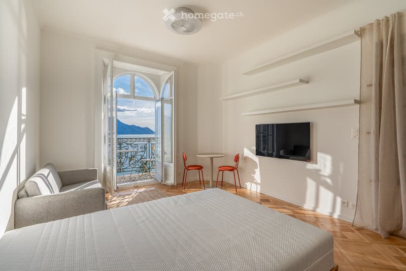 Bright bedroom with a large bed, grey sofa, small round table with two red chairs, wall-mounted TV, built-in shelves, and an open door leading to a balcony with a scenic view.