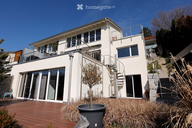 Modern house with wooden deck, white exterior, glass sliding doors, large windows, balcony, spiral staircase, garden, trees, and stairs leading to upper levels