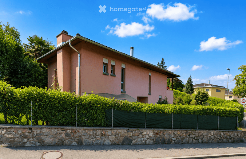 A two-story pink house with a tiled roof, multiple windows, and a chimney, surrounded by greenery and a stone wall with a hedge.
