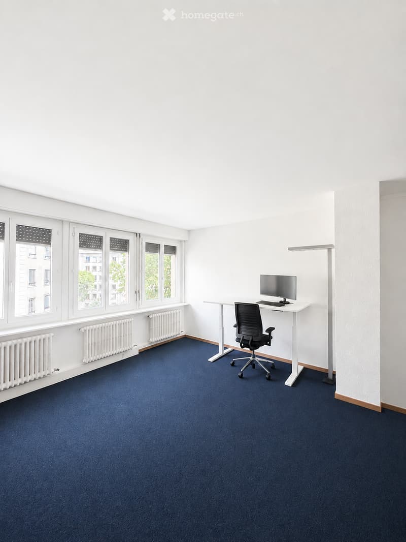 A modern, furnished room with a blue carpet, white walls, large windows, a desk with a computer and screen, and a black office chair.