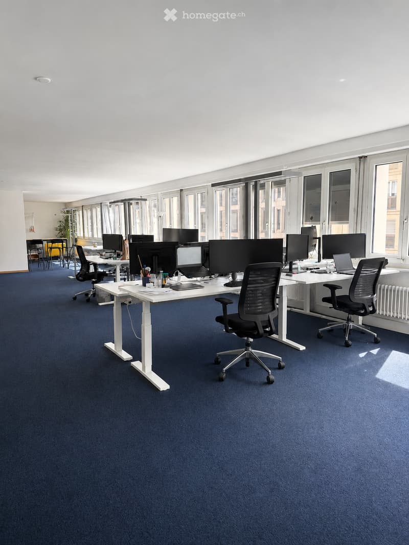 Modern office space with blue carpet, white desks, ergonomic chairs, multiple monitors, laptops, and office supplies.