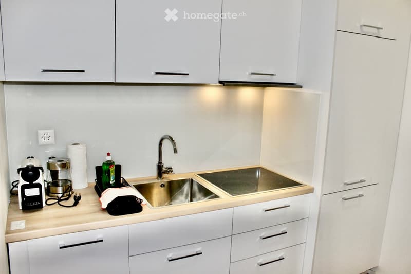 Modern kitchen with white cabinets, stainless steel sink, and countertop. Coffee maker, electric kettle, and cleaning supplies are visible on the counter.