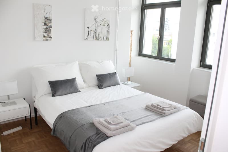 Modern bedroom with a white bed, grey pillows, grey blanket, and neatly folded towels on the bed. Two white nightstands with lamps flank the bed, and a grey stool is placed near the window.