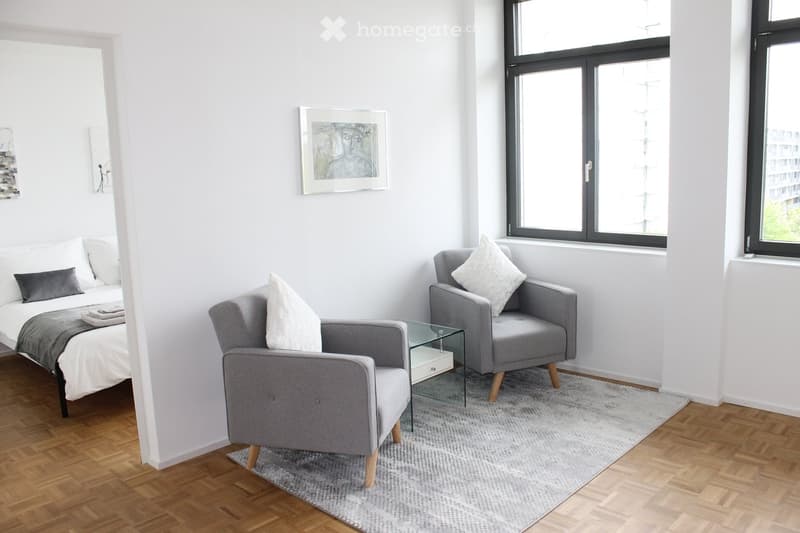 Modern, minimalist bedroom with a bed, two armchairs, a small glass table, and a picture frame on the wall. The room features light-colored walls, wooden flooring, and large windows.