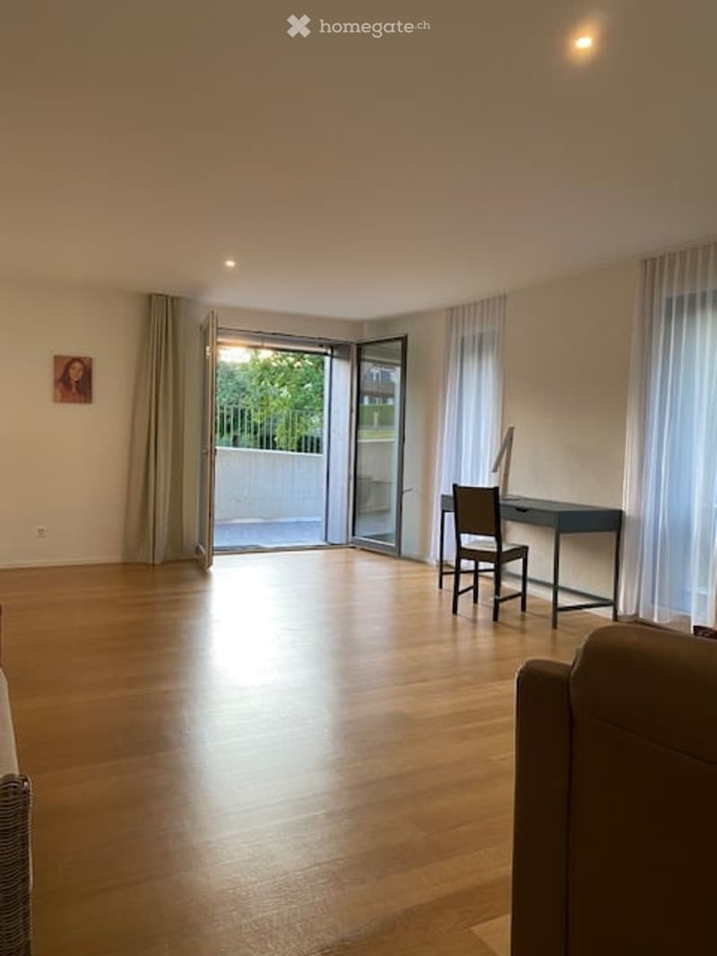 Bright, open living space with wooden flooring, large sliding glass doors leading to a balcony, a sofa, a desk with a chair, and a mirror on the wall.