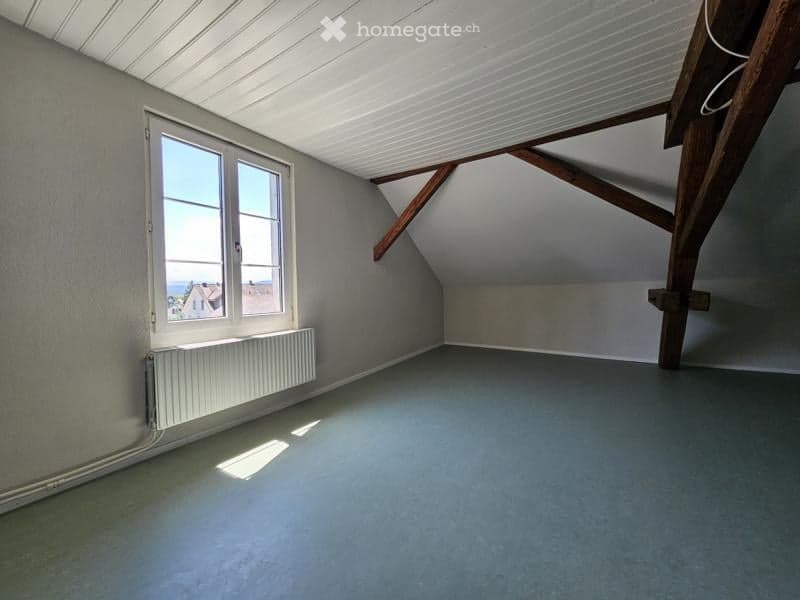 Empty attic room with exposed wooden beams, grey walls, and a large window offering a view of the sea and houses.