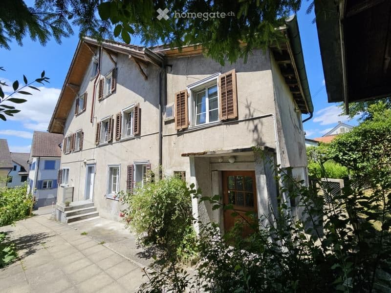 Two-story house with beige facade, wooden shutters, and a small front porch. The house has multiple windows and a front door with glass panels.