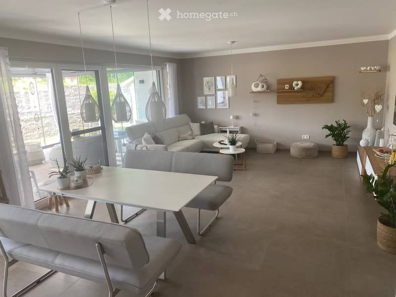 Modern living room with grey sectional sofa, coffee table, dining table, pendant lights, and decorative plants.