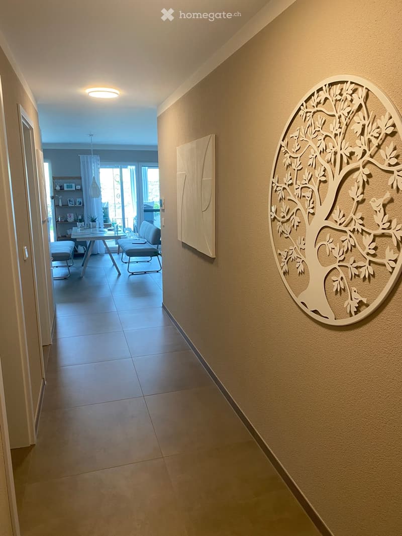 Hallway with beige tiled floor, warm lighting, and a decorative tree wall art piece. The hallway leads to a dining area with a table, chairs, and a view of a balcony.