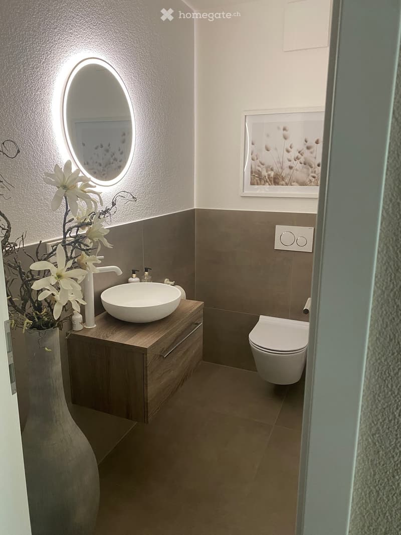 Modern bathroom with a wooden vanity, white sink, oval mirror, toilet, and decorative vase with white flowers.