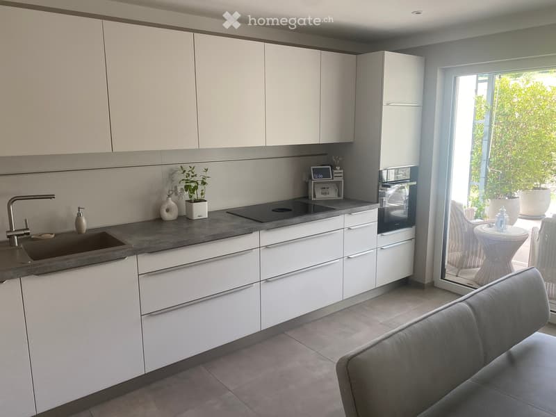 Modern kitchen with white cabinets, grey countertop, sink, faucet, oven, and decorative items including a plant and vase. A grey sofa is partially visible in the foreground.