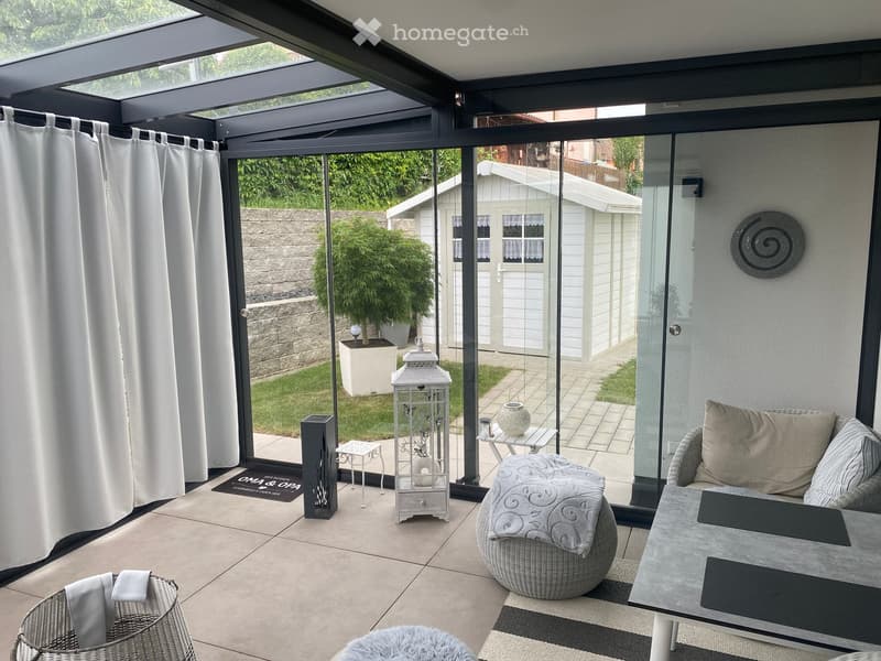 A modern enclosed patio with large glass walls, tiled floor, and comfortable seating including a sofa, coffee table, wicker chairs, and a lantern.