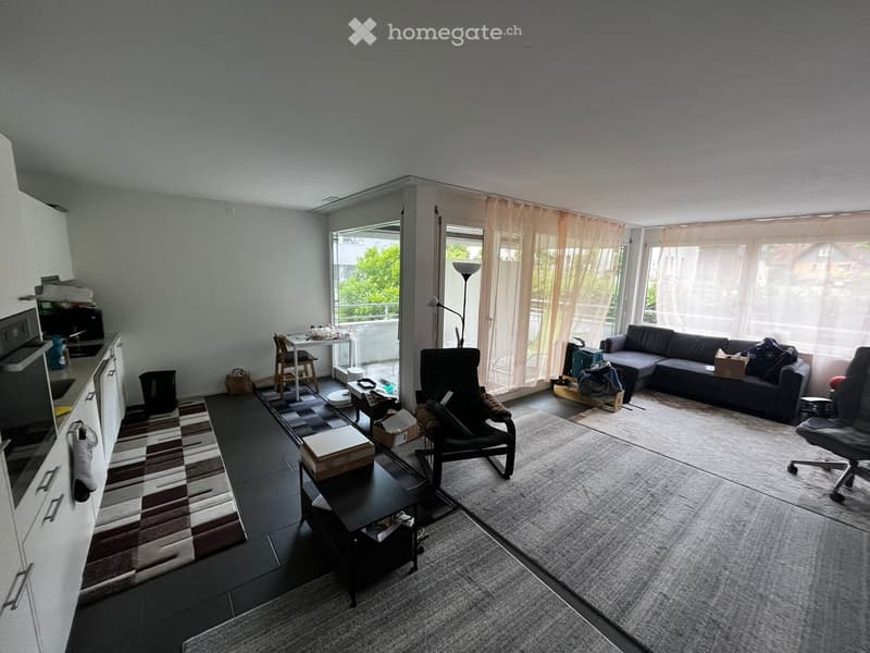 Modern living room with a sofa, armchair, coffee table, kitchen area, and large windows with sheer curtains.