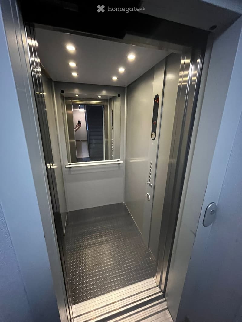 Elevator shaft with metallic walls, control panel, and illuminated ceiling lights. The elevator doors are open, revealing a clean, modern interior with a textured floor and handrail.