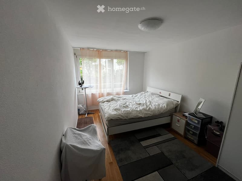 Cozy bedroom with a bed, a covered chair, a small table near the window, a mirror, and storage units.
