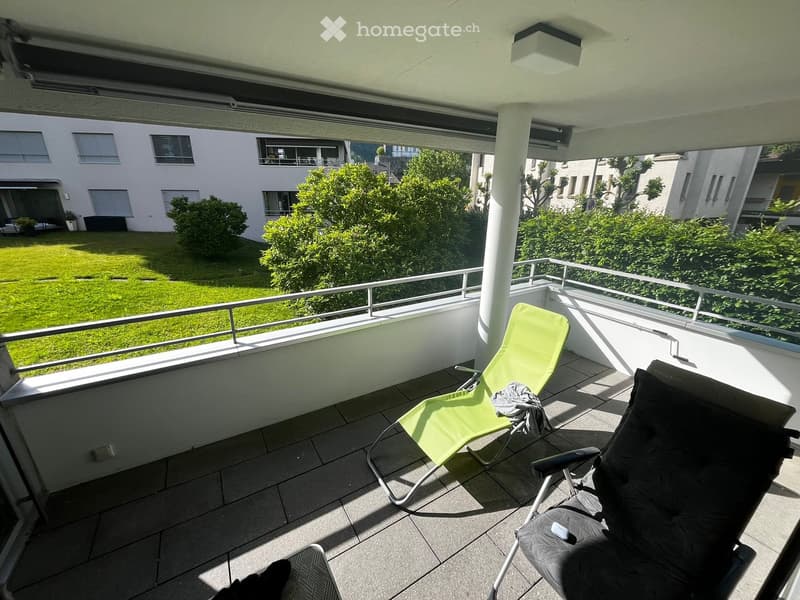 Balcony with tiled floor, metal railing, two chairs, and a view of a green lawn and surrounding buildings.