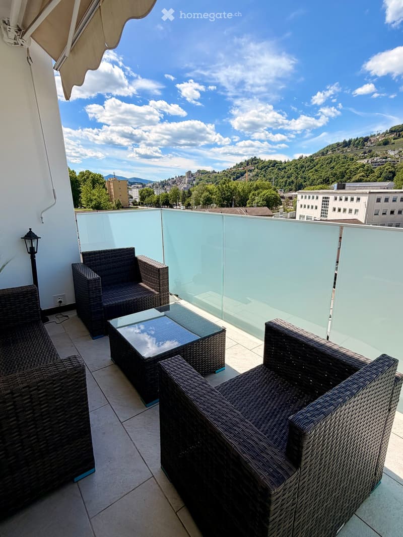Balcony with wicker chairs and a glass table, overlooking a cityscape with trees and buildings.