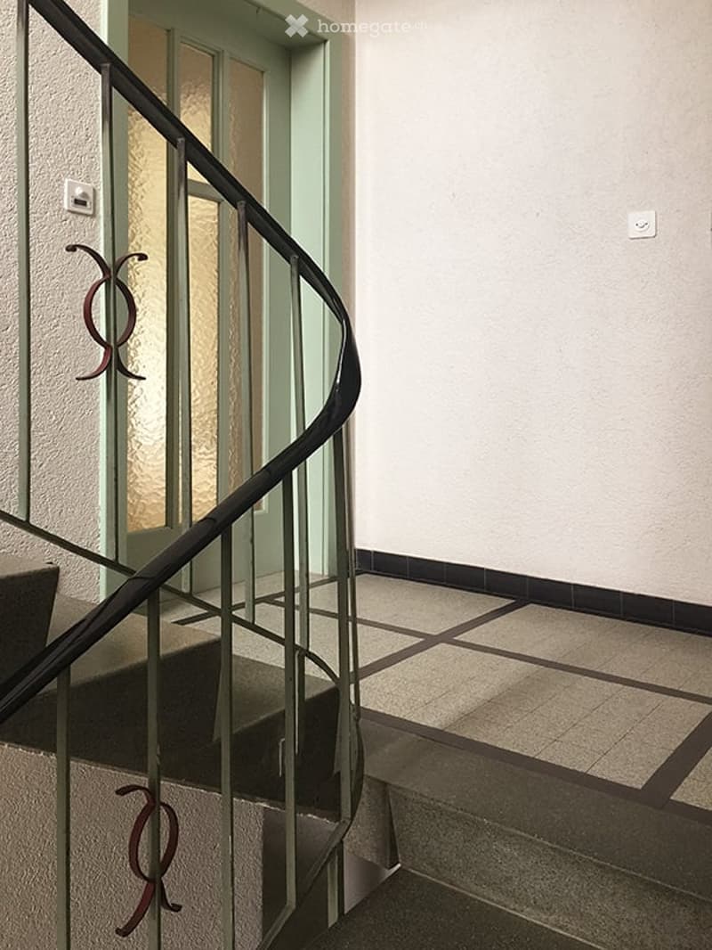 Spacious hallway with a modern staircase featuring a black metal railing and glass panels. The walls are painted white, and the floor is tiled with a geometric pattern.