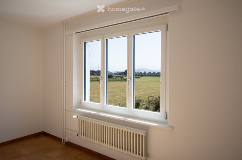 Empty room with wooden flooring, white walls, large windows with white frames, and a radiator below the windows.