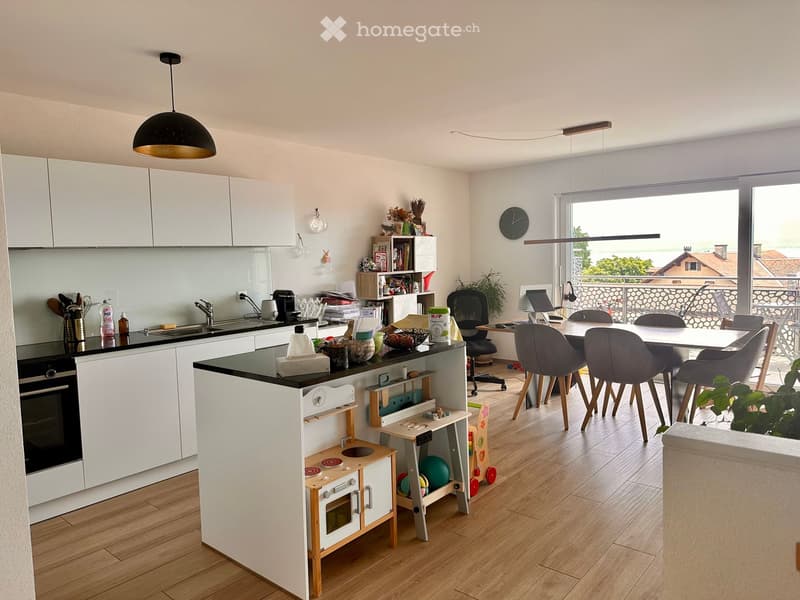 Modern kitchen with white cabinets, black countertop, and a small play area for children. There is a dining area with a table and chairs, and a laptop on a desk near a large window with a view.