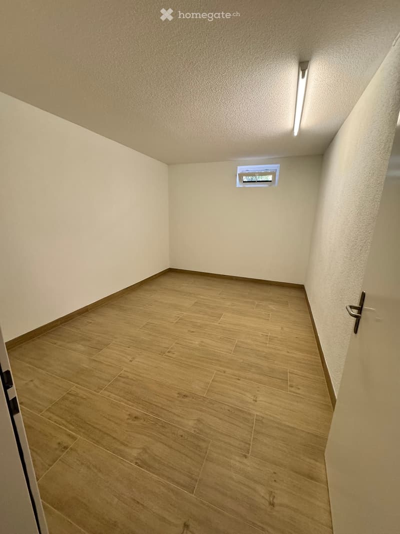Empty hallway with light wooden flooring, white walls, and a small window at the far end.