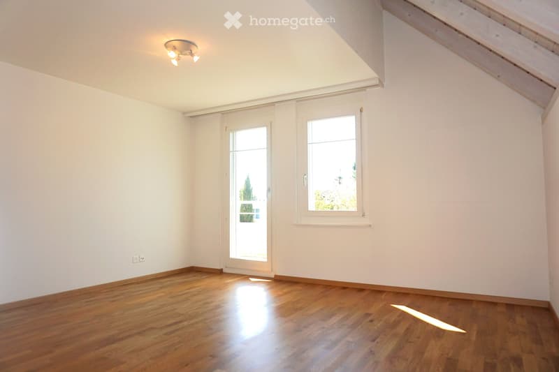 Empty room with wooden flooring, white walls, and large windows allowing natural light. The room features a sliding glass door leading outside and a ceiling light fixture.