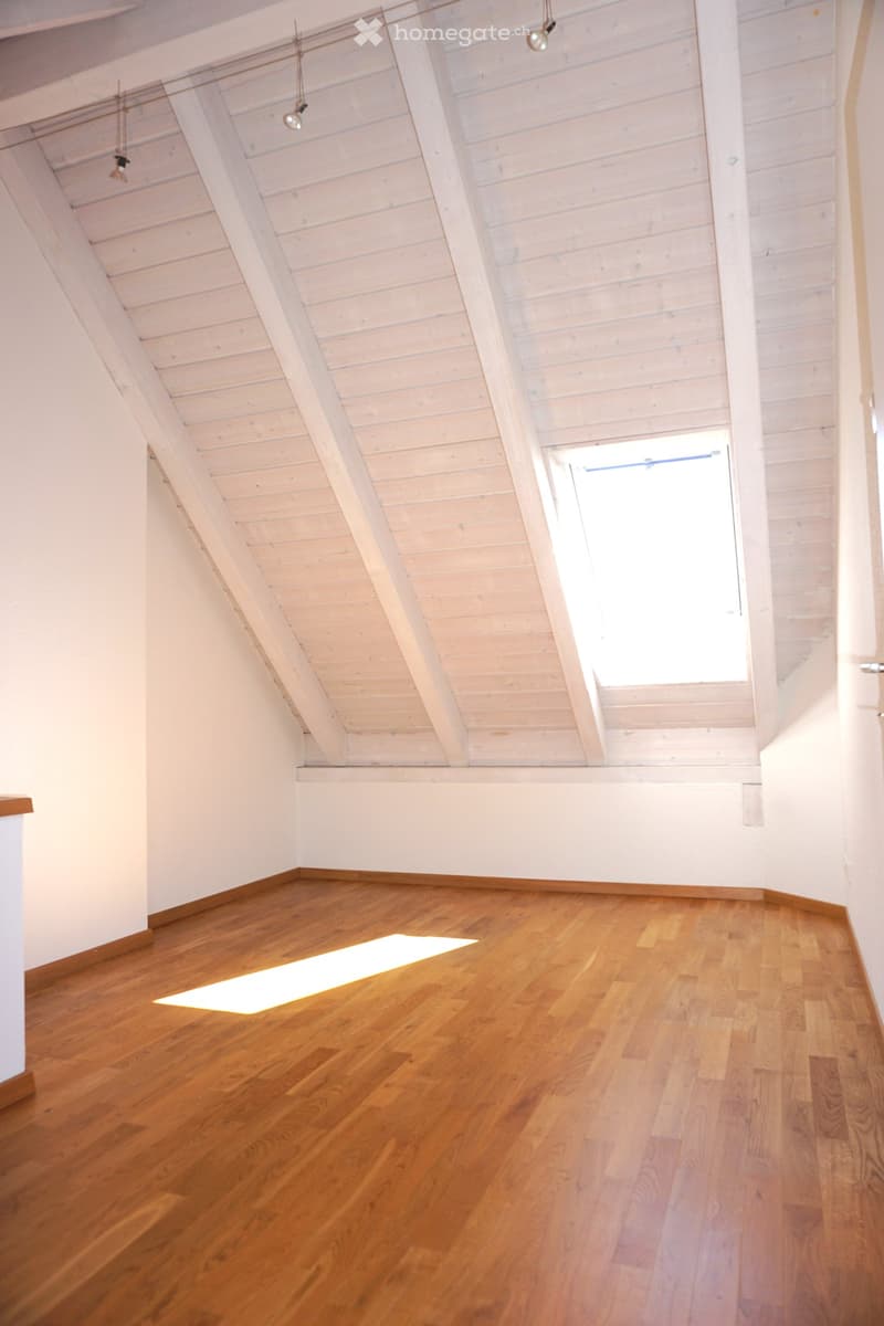 Empty attic room with wooden floors, white walls, and exposed wooden ceiling beams. A skylight window allows natural light to enter.