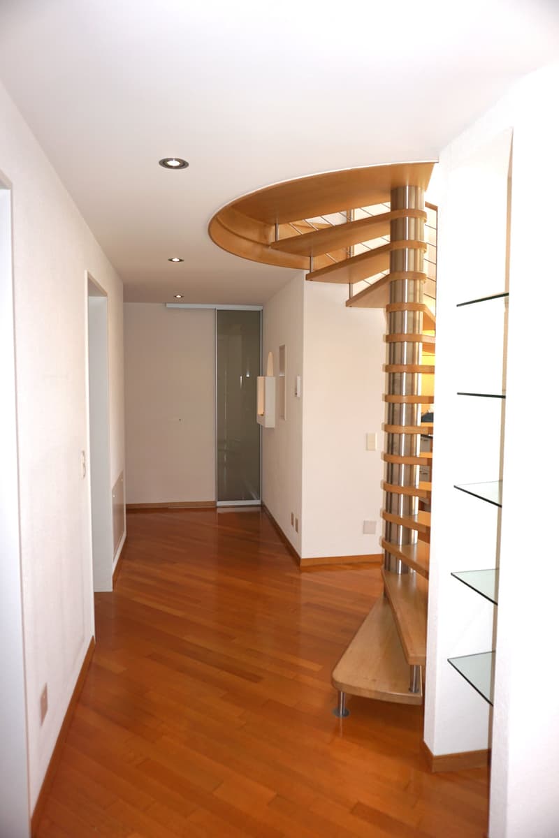 Modern hallway with wooden flooring, white walls, recessed lighting, and a curved staircase with glass railings leading to another level.