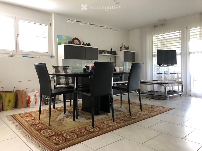 Modern dining area with a black table, four black chairs, a cabinet, and a television on a stand. The room features large windows with white curtains and a patterned rug on the floor.