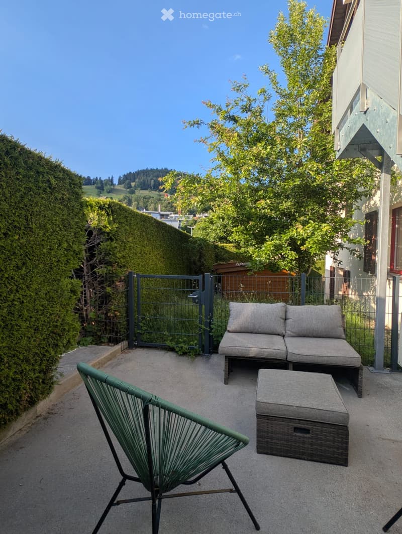 A small outdoor patio with a grey sofa, a green chair, and a small table, surrounded by greenery and a metal gate.