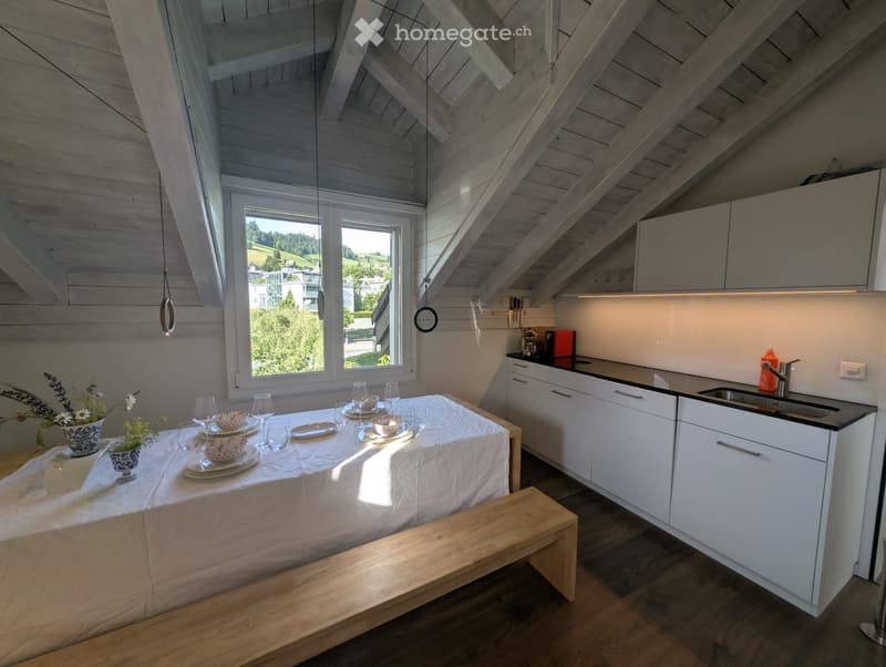 Bright, airy room with exposed wooden beams, a dining table set for four, a modern kitchen area with white cabinets and black countertops, and a view of greenery through large windows.